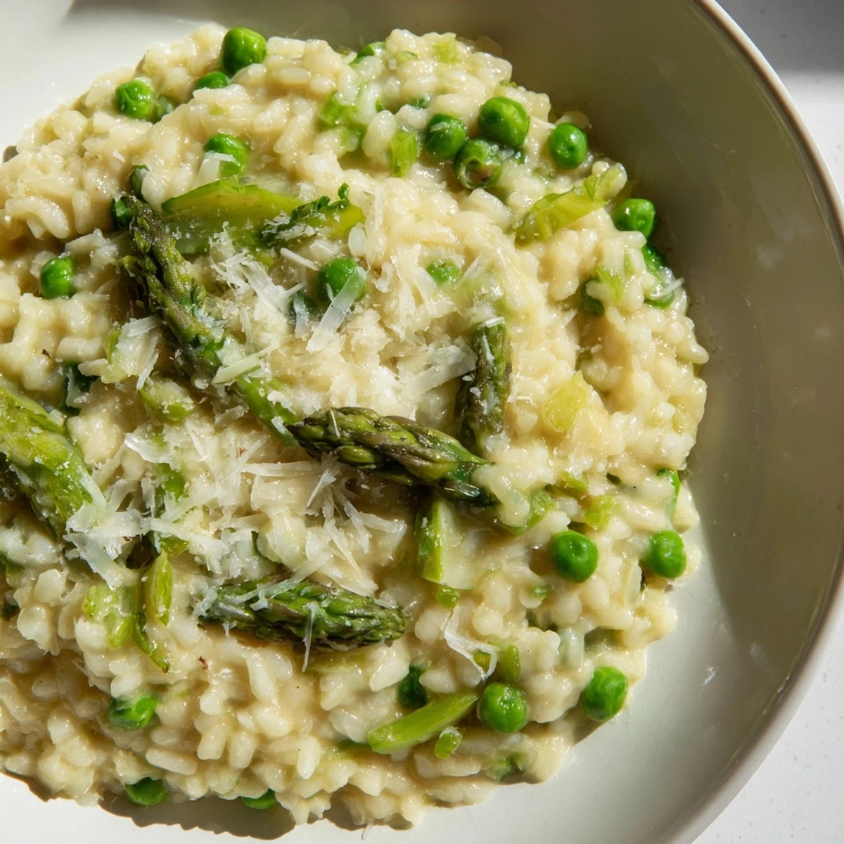 A close-up of creamy asparagus and pea risotto garnished with fresh Parmesan in a rustic white bowl.