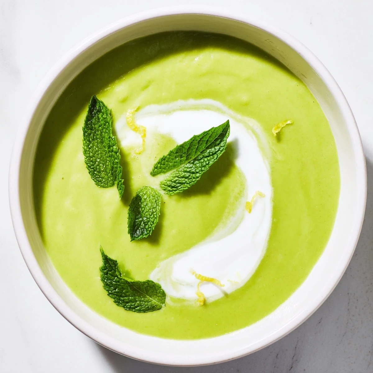 Top-down view of Creamy Spring Pea and Mint Soup with Crème Fraîche, showing a decorative swirl of tangy crème fraîche and a lemon zest garnish.