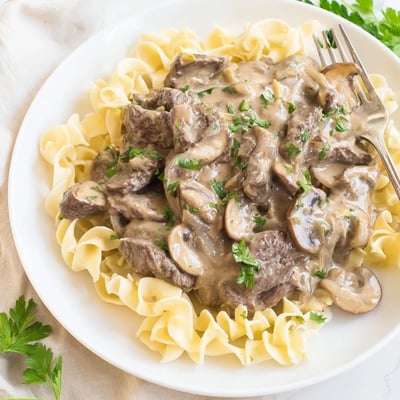 Golden beef stroganoff plated over noodles with fresh parsley garnish on white background